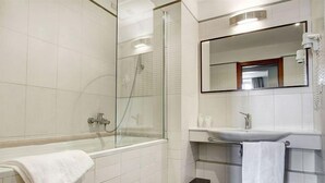 Un baño moderno con ducha de vidrio, un lavamanos blanco con espejo y una toalla blanca sobre el piso.