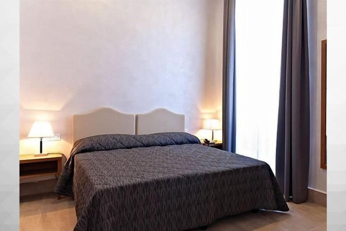 A hotel room with a bed, two bedside lamps, and a window with curtains.