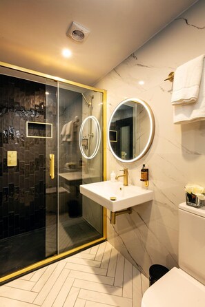 Uma casa de banho moderna com chuveiro em cabine de vidro, pia branca com detalhes dourados e espelho redondo.