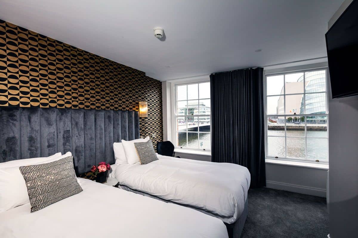 A hotel room with two beds, a patterned headboard, a large window with a view of a body of water and buildings, and a flat-screen TV mounted on the wall.