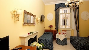 A hotel room with two single beds, a desk with a chair, a television, a fireplace, and a chandelier. - Caroline Suite (Rome)