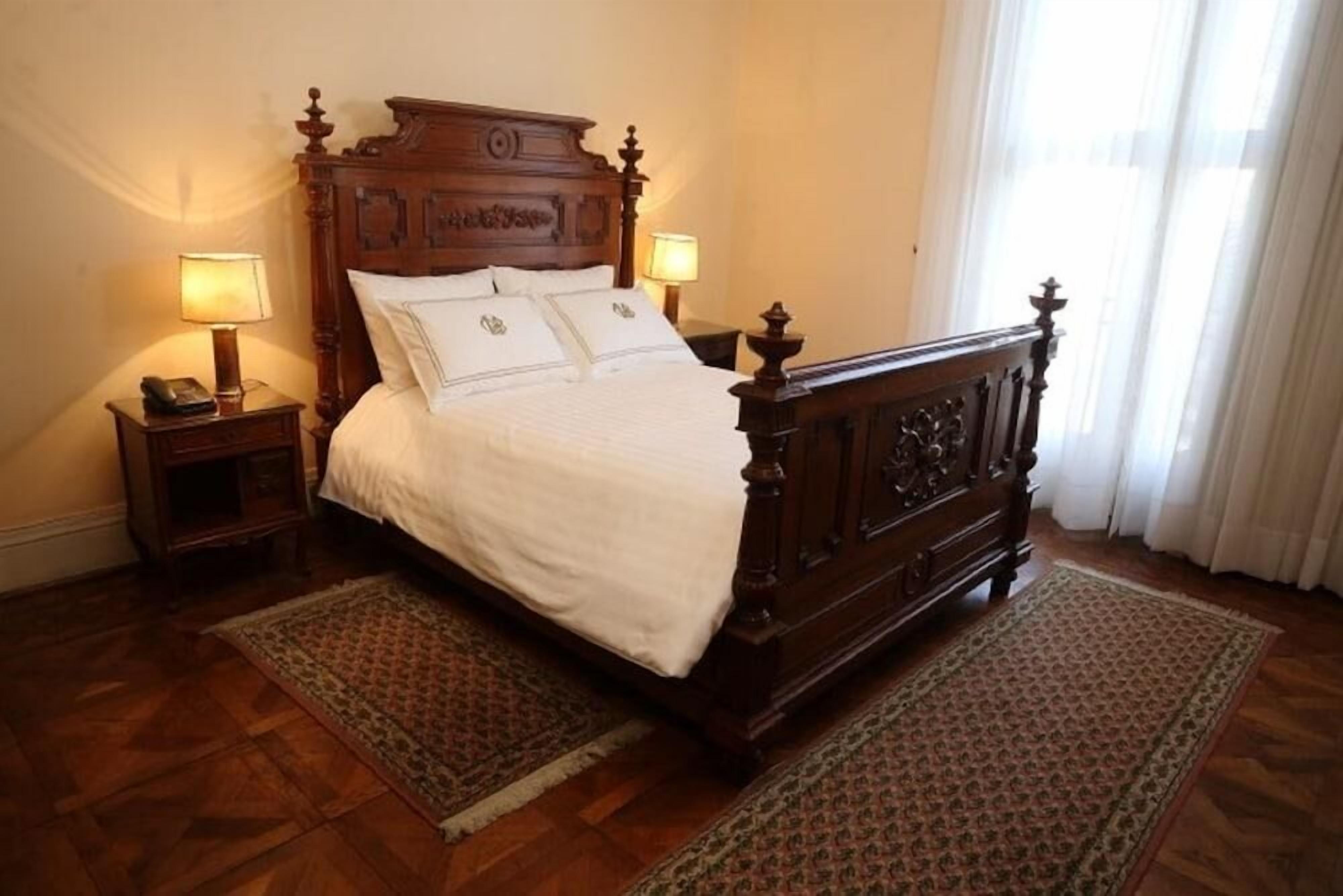 A traditional wooden bed with a headboard, two bedside tables with lamps, and patterned rugs on a wooden floor.