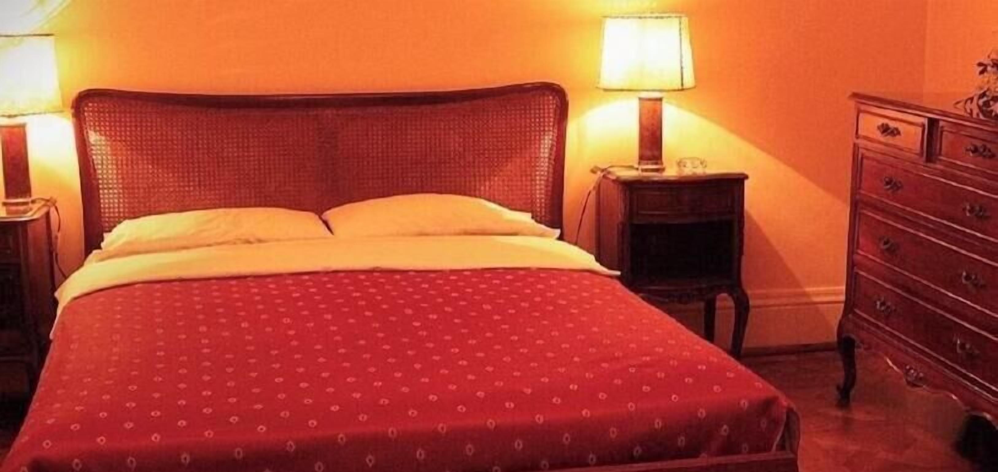 A bed with a patterned bedspread, a nightstand with a lamp, and a dresser with drawers.