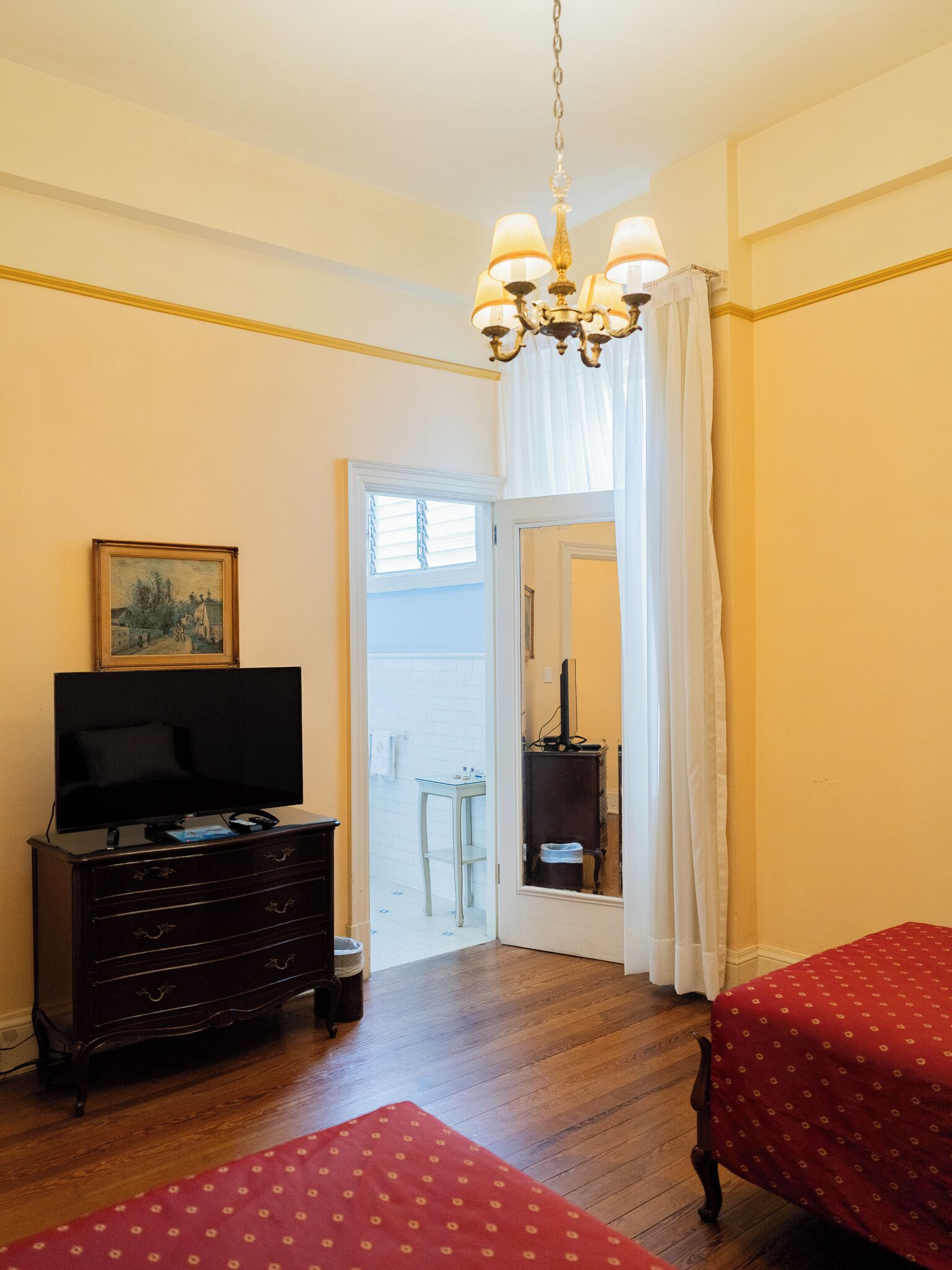 Una habitación con televisión sobre una cómoda, una cama con cubrecolcha rojo, una araña y una puerta que da a un balcón.