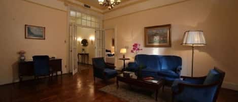 A room with a blue sofa, two armchairs, a coffee table, and a lamp. There is a door leading to another room.