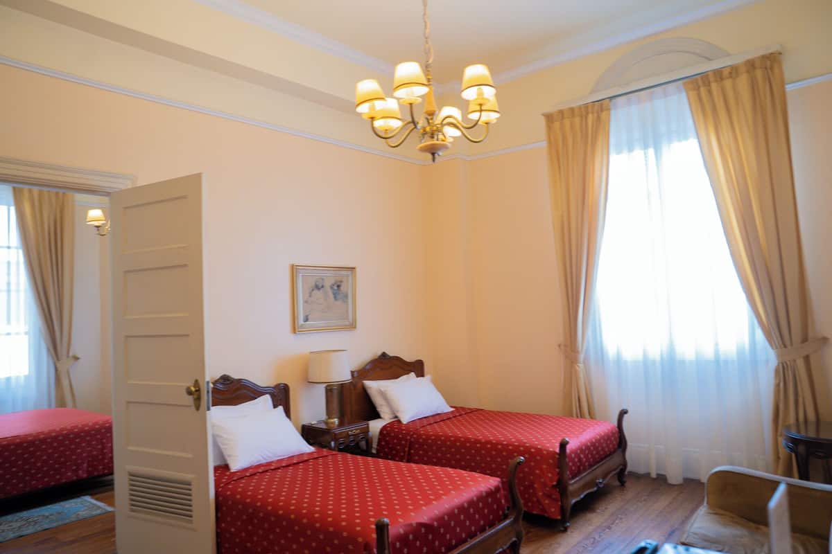 A hotel room with two beds, a chandelier, a painting on the wall, and a window with curtains.