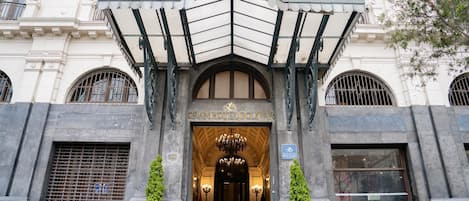 A entrada do Gran Hotel Bolivar, com estilo arquitetônico clássico, apresenta um portal imponente, um lustre e duas plantas em vasos.