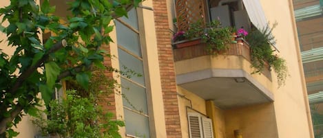 A building with multiple balconies, some with plants and flowers. There are visible windows and a door, and the building has a brick facade.