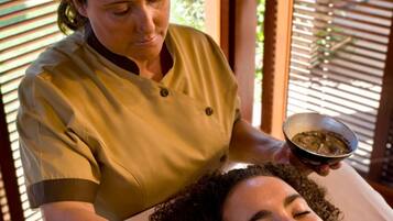 A person receiving a facial treatment in a room with wooden blinds and a view of greenery outside.