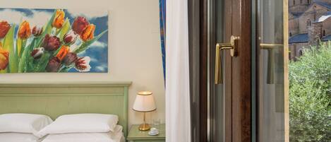 Habitación de hotel con cama, mesita de noche con lámpara, un cuadro de tulipanes y ventana con vista a un edificio.