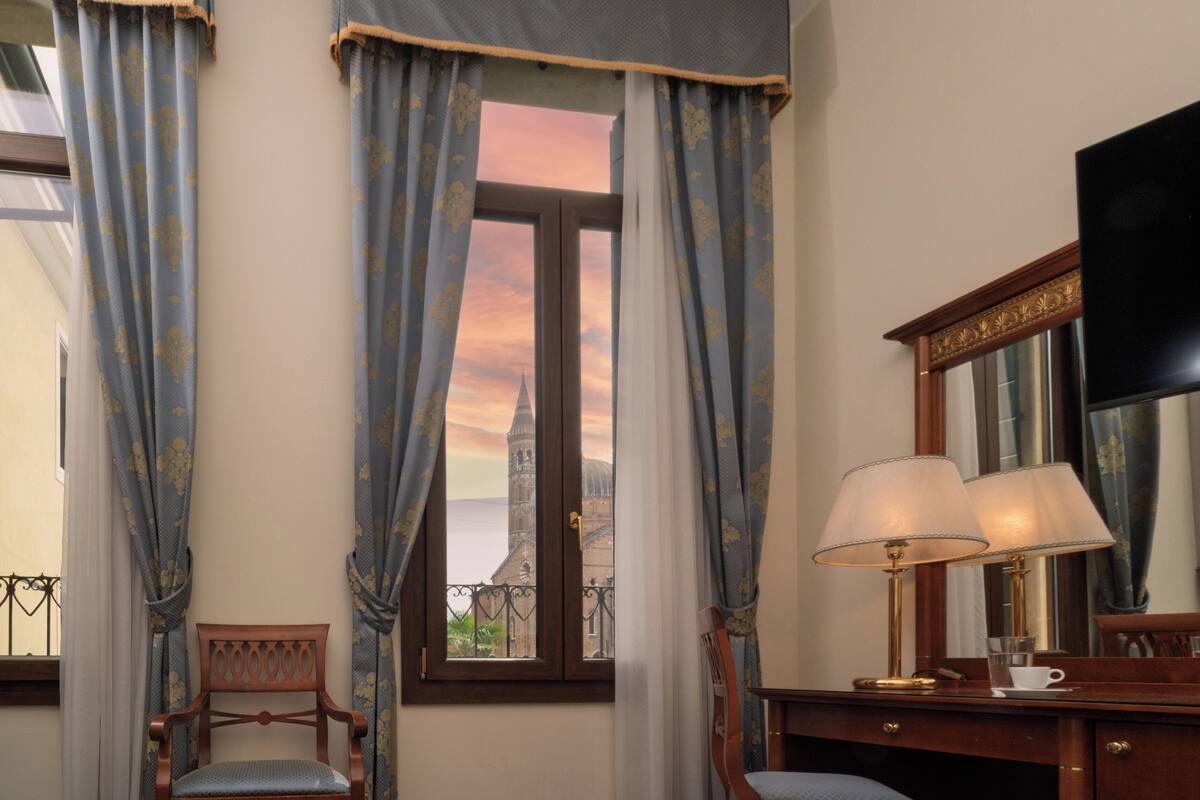 Habitación de hotel con un ventanal grande, cortinas, un escritorio de madera con lámpara, una silla y un televisor de pantalla plana.
