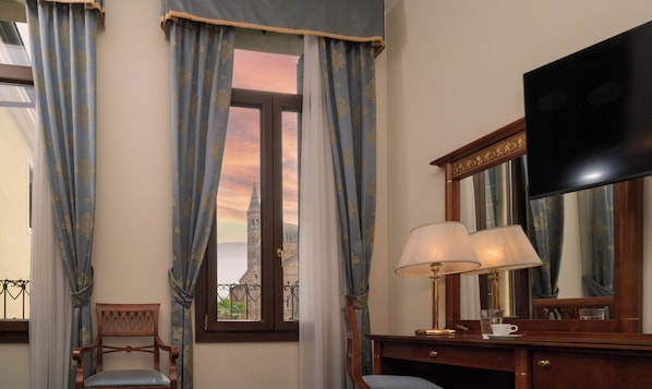 A hotel room with a large window, curtains, a wooden desk with a lamp, a chair, and a flat-screen TV. - Donatello (Padova)