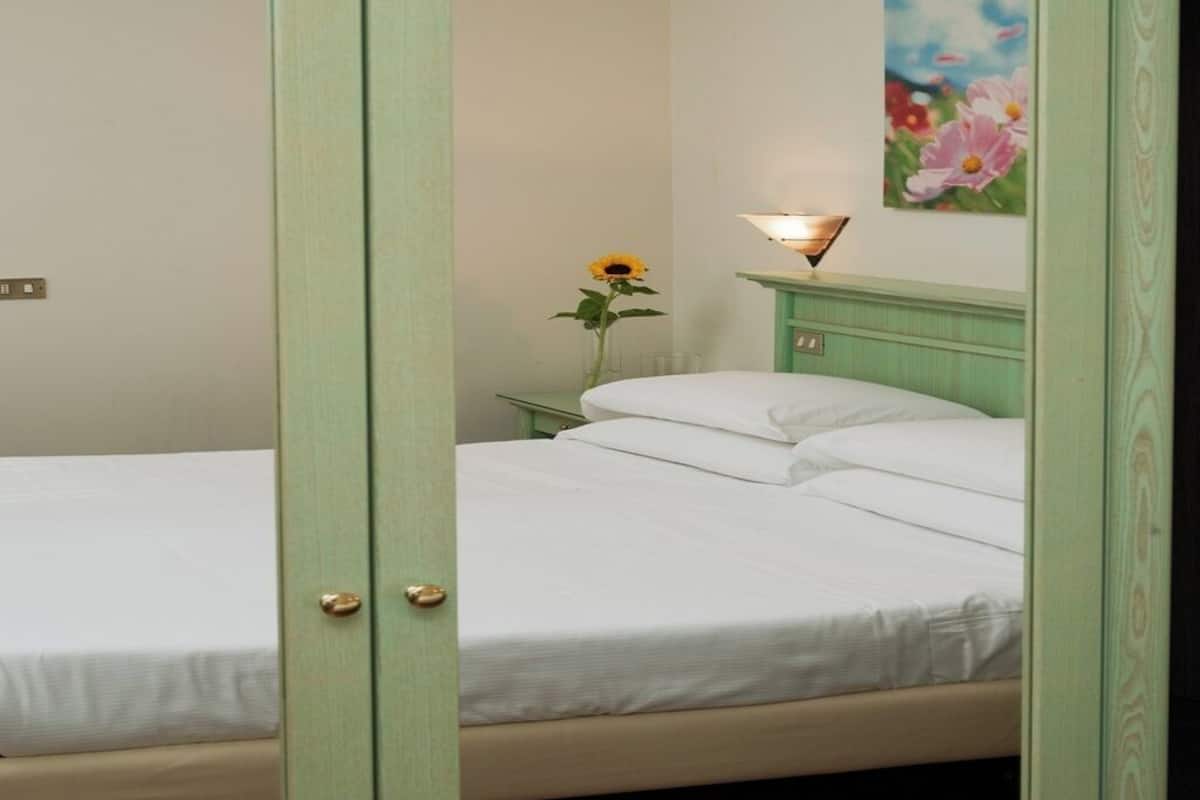 Una habitación con dos camas, una mesita de noche con una lámpara, un cuadro en la pared y un jarrón con un girasol.