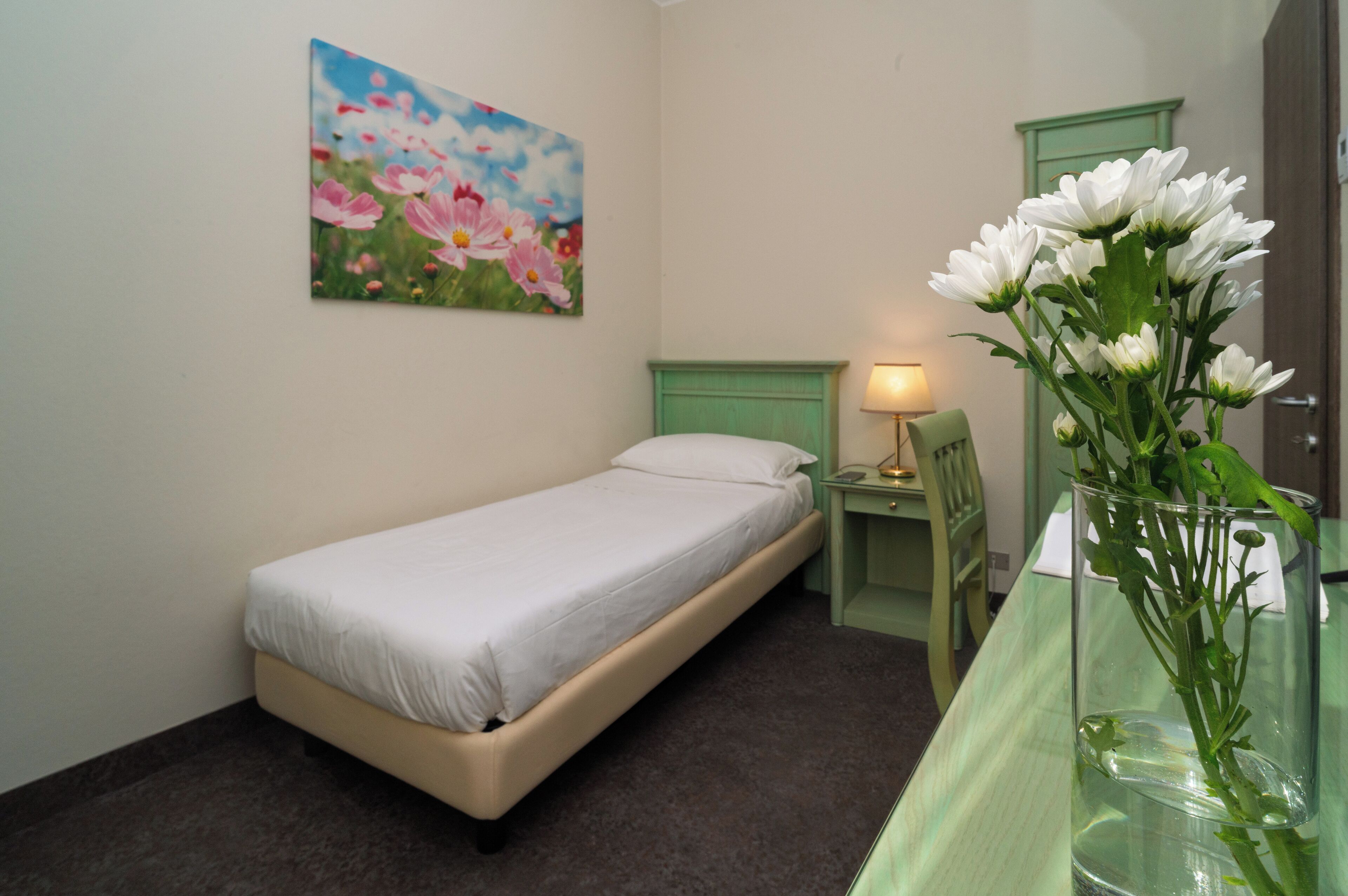 A small, single-bed room with a green headboard, a bedside table with a lamp, and a vase with flowers on a glass table.