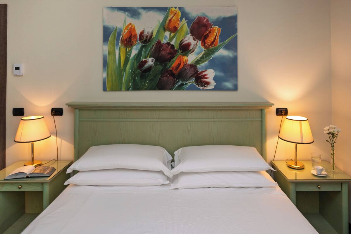 Una cama bien hecha con sábanas blancas, dos mesitas de noche con lámparas, un cuadro de tulipanes en la pared y un pequeño florero con una sola flor en una de las mesitas.