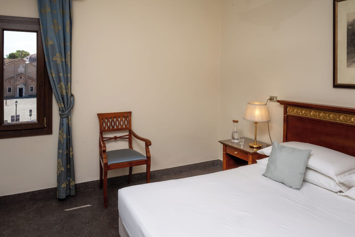 Habitación de hotel con cama, una silla de madera, mesita de noche con lámpara y ventana con cortinas.