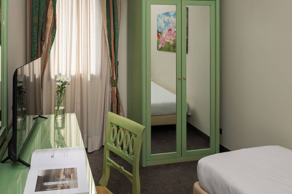 Una habitación con cama, un armario verde, un espejo, una mesa con un jarrón de flores y una ventana con cortinas traslúcidas.