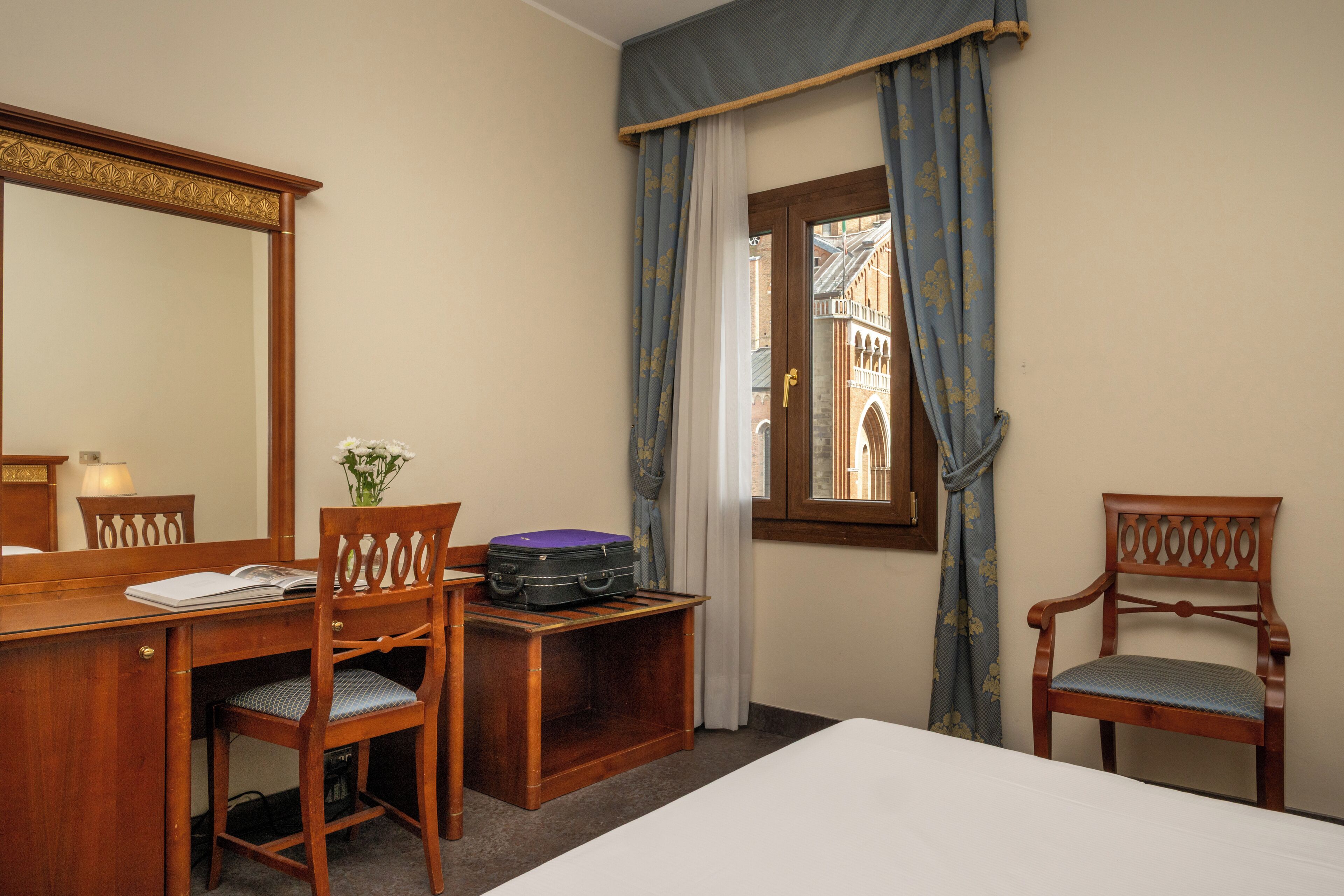 A hotel room with a wooden desk, two chairs, a bed, a window with curtains, and a mirror.