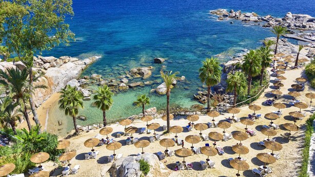 A beach with clear blue water, rocky shore, palm trees, and sun loungers with umbrellas. - Arbatax Park Resort - Suites del Mare by Falkensteiner (Tortoli)