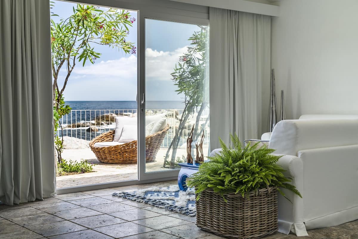 Uma sala de estar com um sofá branco, uma cesta de vime com plantas verdes e vista para o mar através de uma porta de vidro.