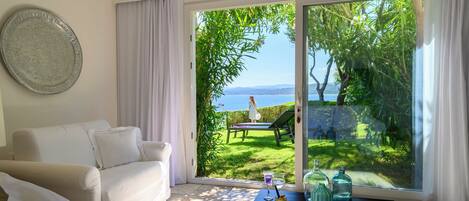 A room with a white sofa, a glass table with bottles, and a view of a garden and sea through large glass doors.