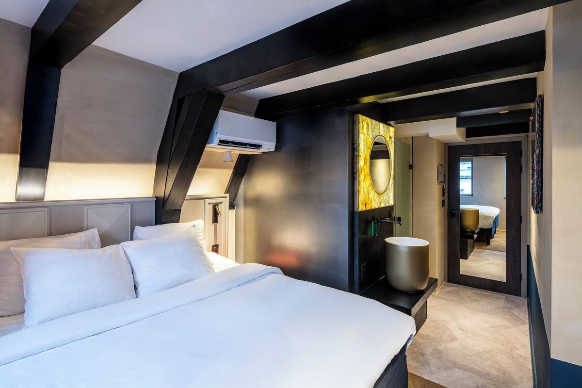 Una habitación de hotel moderna con una cama grande, un baño con un espejo amplio y vista a la habitación contigua.
