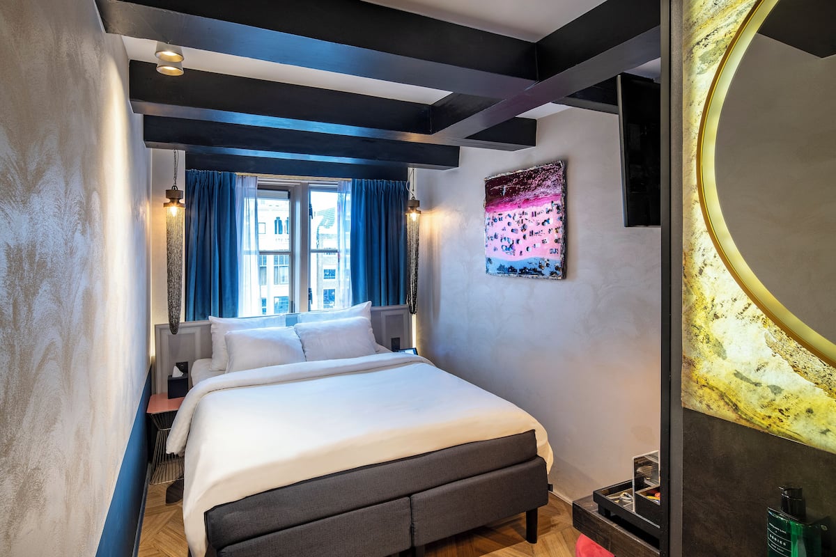 Una habitación de hotel moderna con una cama grande, una ventana con cortinas azules, una obra de arte colgada en la pared y un espejo redondo.