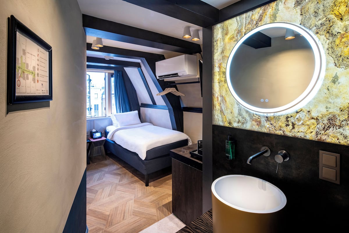 Habitación de hotel moderna con una cama, un espejo redondo, un lavamanos y vistas de la ciudad a través de una ventana.