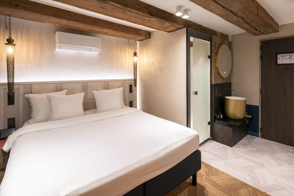 Un dormitorio con una cama grande, vigas de madera, aire acondicionado de pared y un baño con un espejo grande y lavamanos.