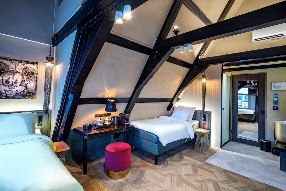 Un dormitorio tipo loft moderno con vigas a la vista, una cama grande, una cama más pequeña, un escritorio y un reposapiés rojo.