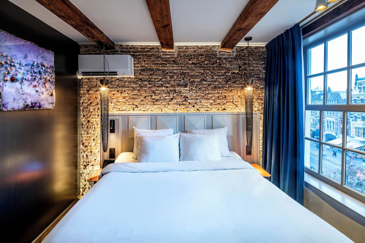 Habitación de hotel con pared de ladrillo, una cama grande con ropa blanca, cabecero con lámpara y vista a la ciudad por la ventana.