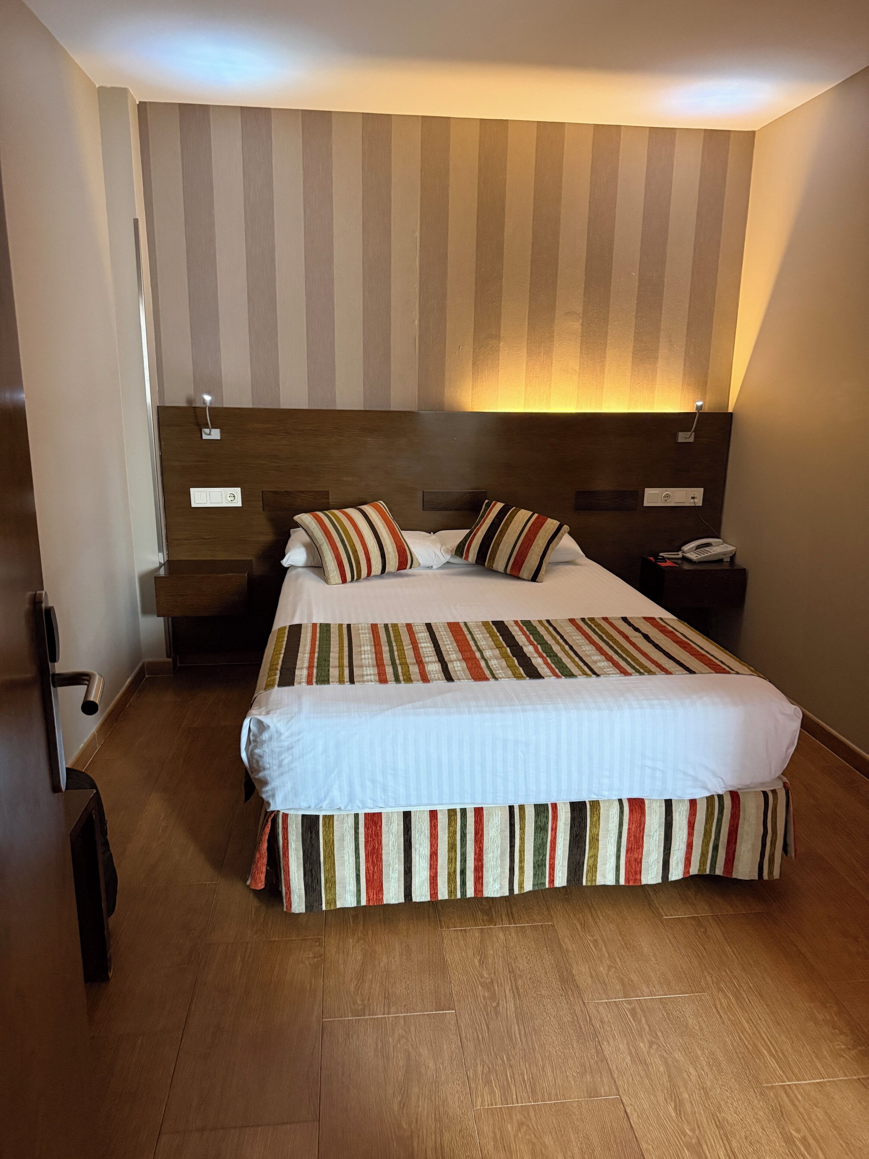 Una habitación de hotel con una cama grande, un cabecero de madera, una mesita de noche con un teléfono y una lámpara fijada a la pared.