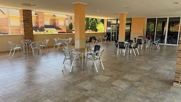 Un patio al aire libre con mesas y sillas de metal, piso de baldosas y vista a edificios en el fondo.