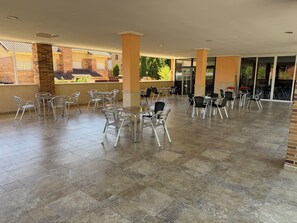 Un patio al aire libre con mesas y sillas de metal, piso de baldosas y vista a edificios en el fondo.