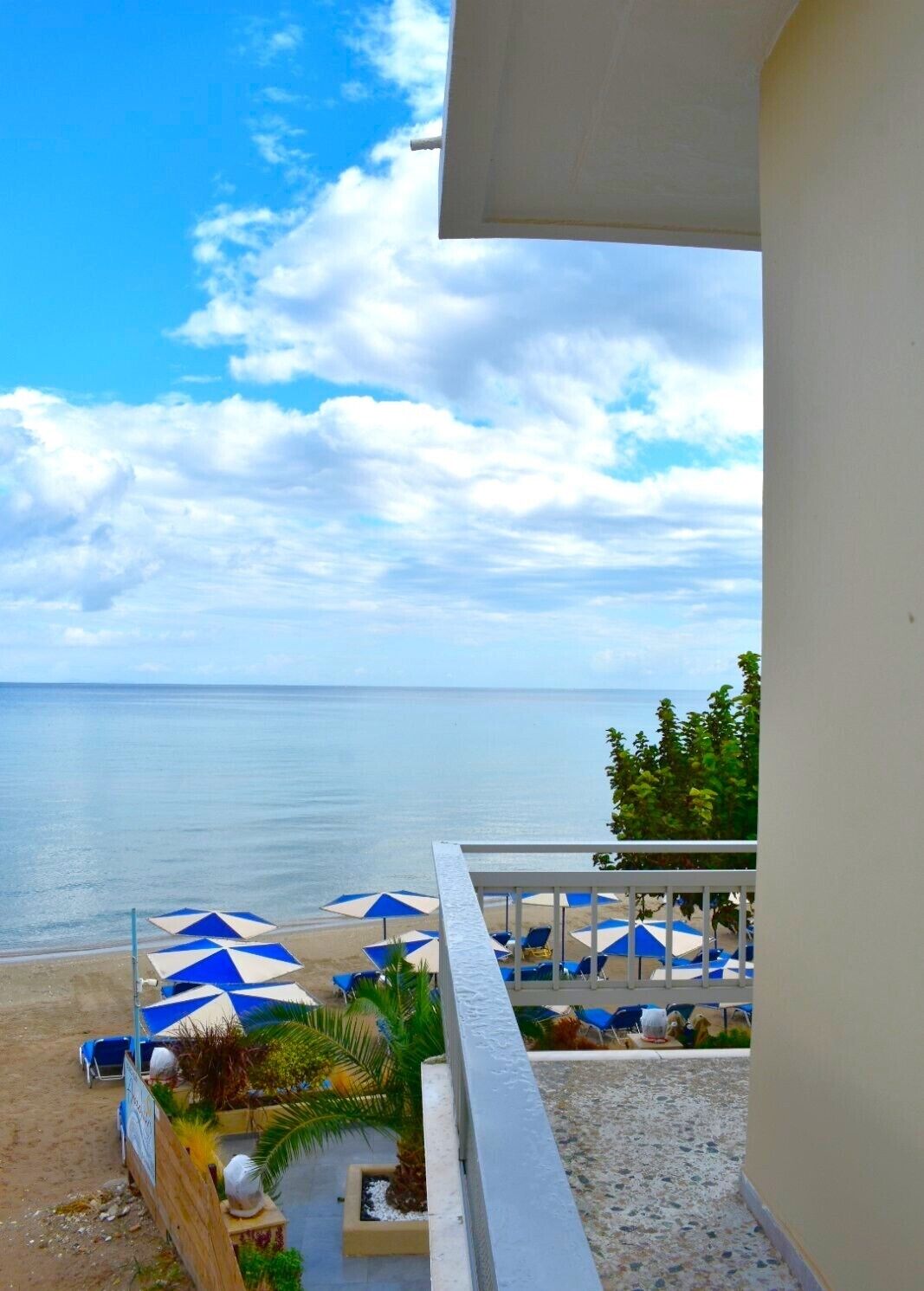 Un balcon avec vue sur la plage, agrémenté de parasols à rayures bleues et blanches, de chaises longues et d’une zone sablonneuse.