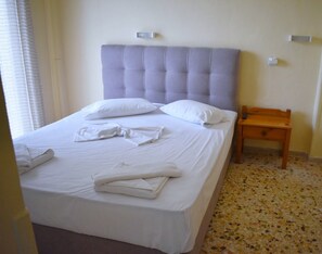 A neatly made bed with white linens and pillows, a wooden nightstand with a drawer, and a tiled floor. - Asteria (Zakynthos)