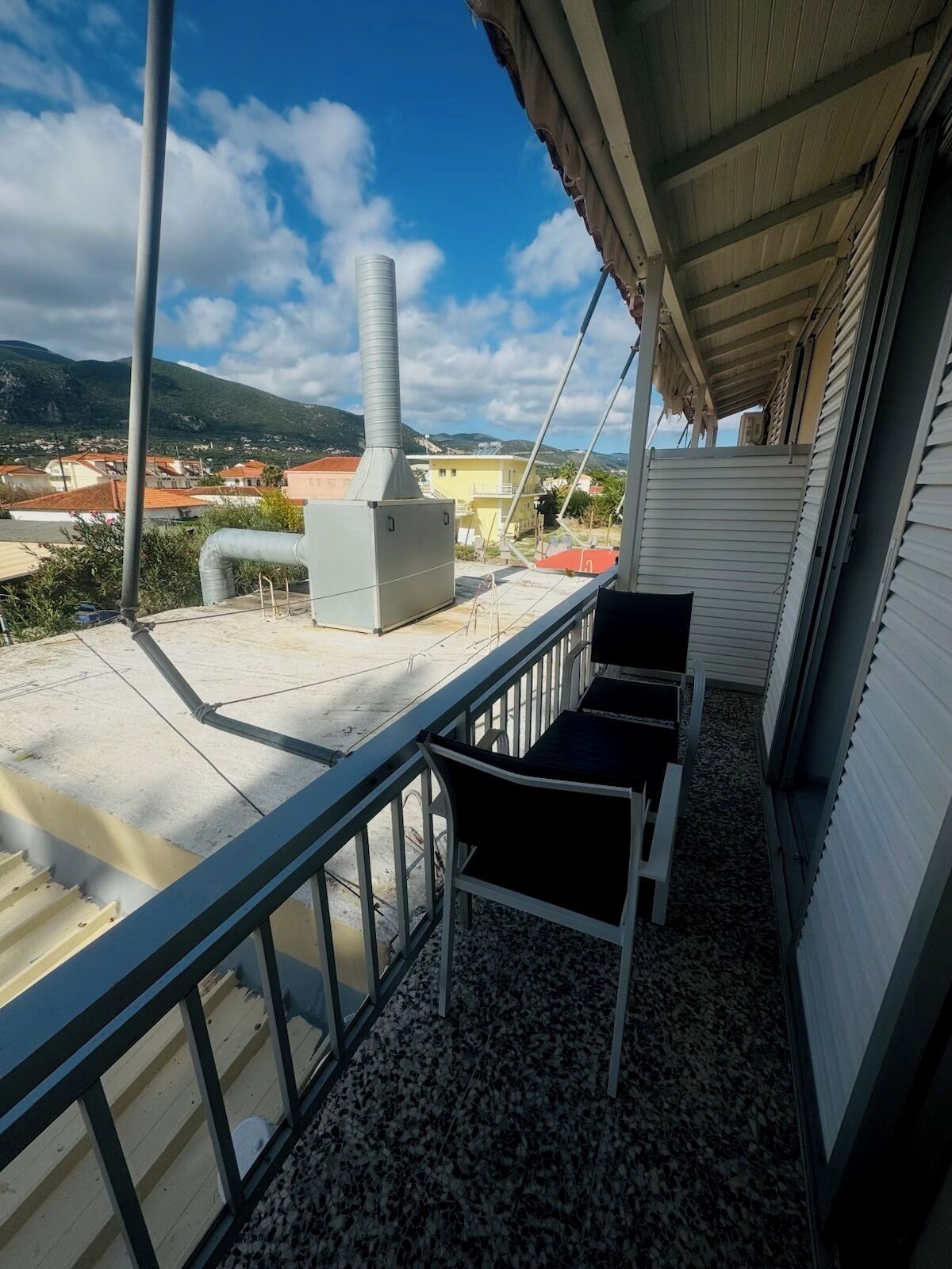 Un balcon avec une balustrade en métal, une chaise blanche et une chaise noire. On aperçoit, en arrière-plan, un bâtiment blanc avec un toit rouge.