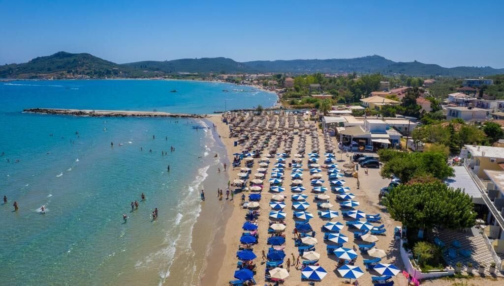 Une plage parsemée de nombreux parasols, une mer d’un bleu limpide et une ville qui se dessine à l’arrière-plan.