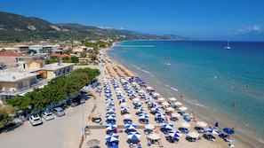 A coastal town with a beach lined with umbrellas and sun loungers, a clear blue sea, and buildings along the shore.