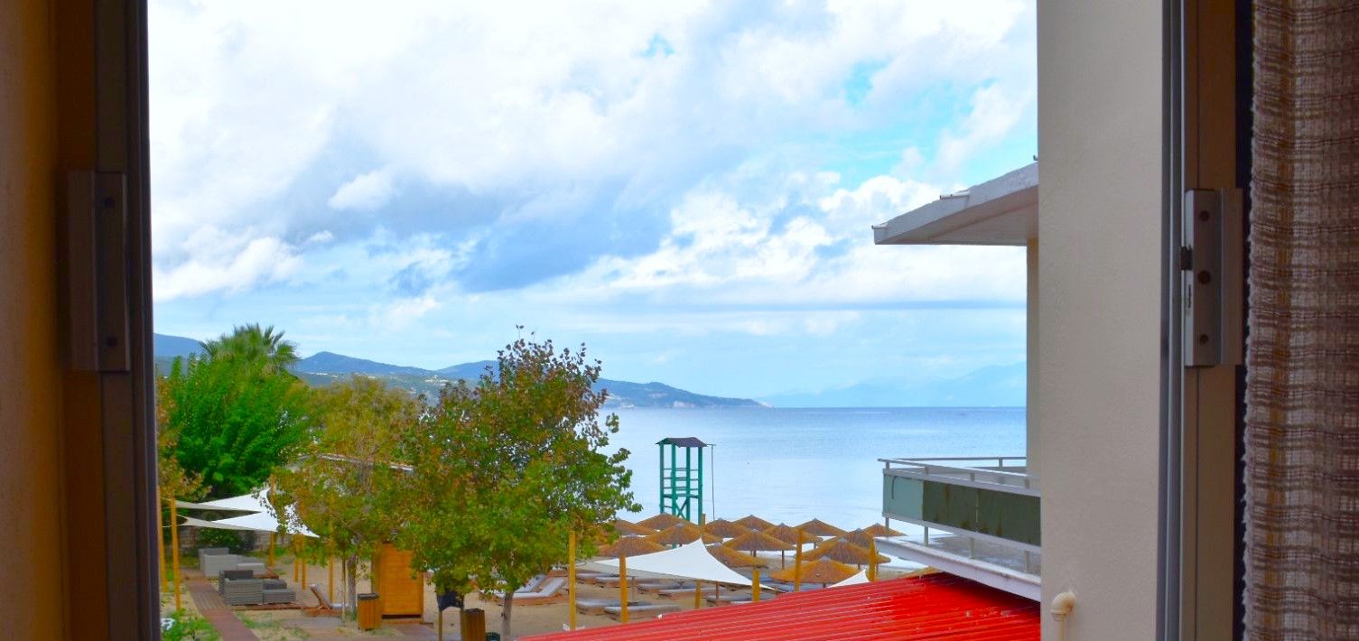 Vista da una stanza con finestra su una spiaggia, una torretta di salvataggio e un edificio con tetto rosso.