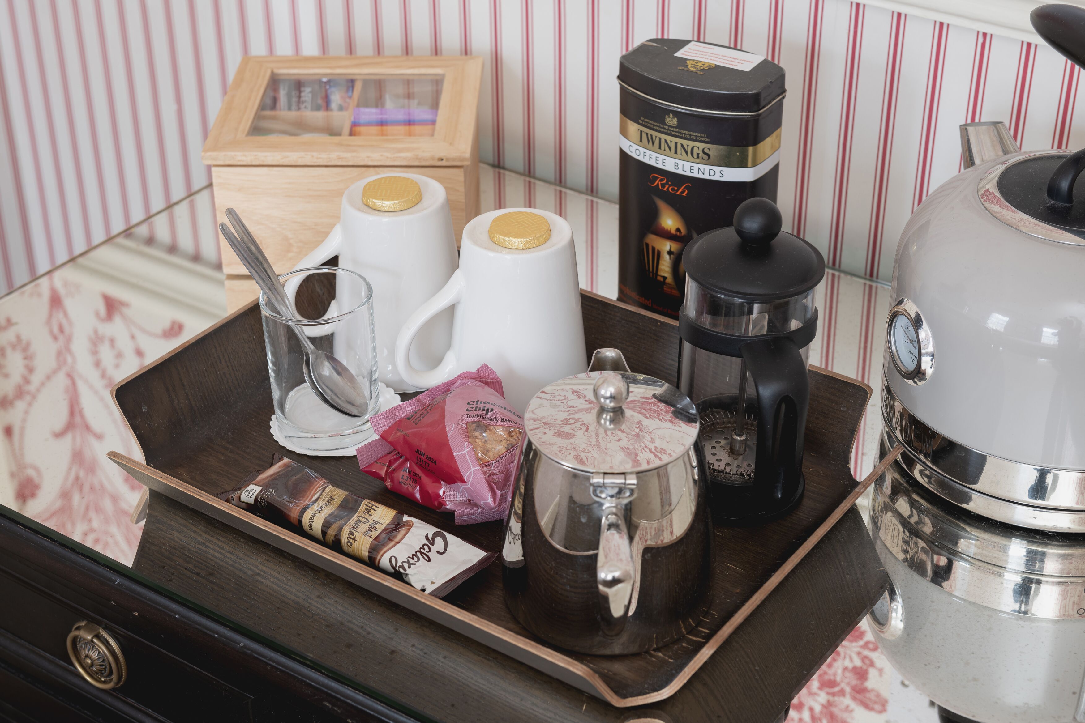 A tray with a coffee maker, two cups, a glass, a packet of snacks, and a box of Twinings coffee blends.