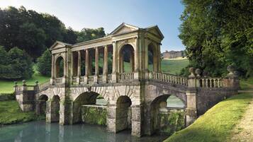 A stone bridge with arches over a calm river, surrounded by lush greenery and a distant building.