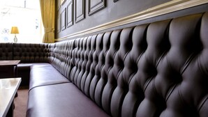 A dark-colored, tufted bench with a high backrest, positioned against a wall with framed panels and a decorative molding. - The Royal Hotel (Bath)