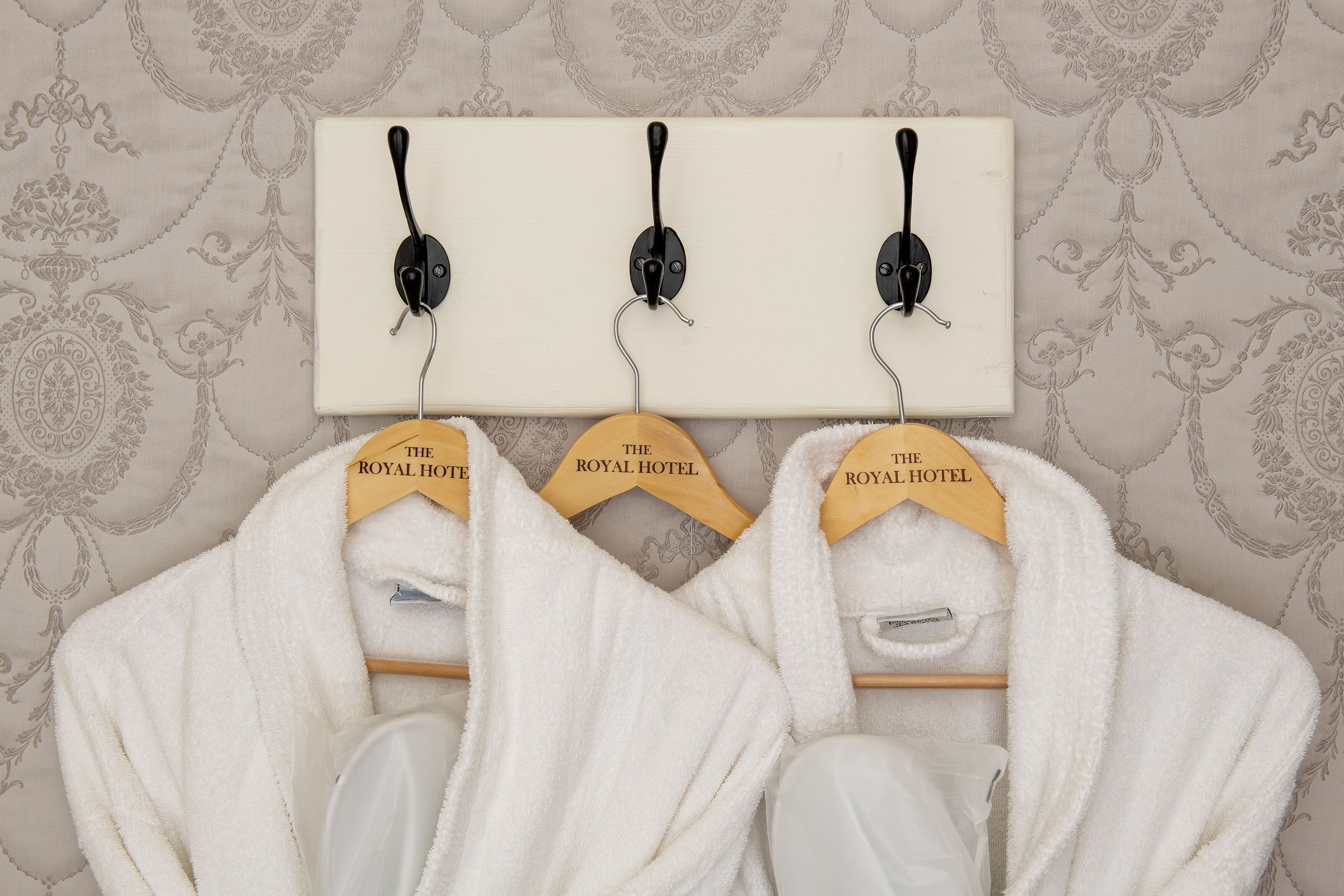 Three white robes hanging on wooden hangers labeled "The Royal Hotel" on a wall with a patterned wallpaper background.
