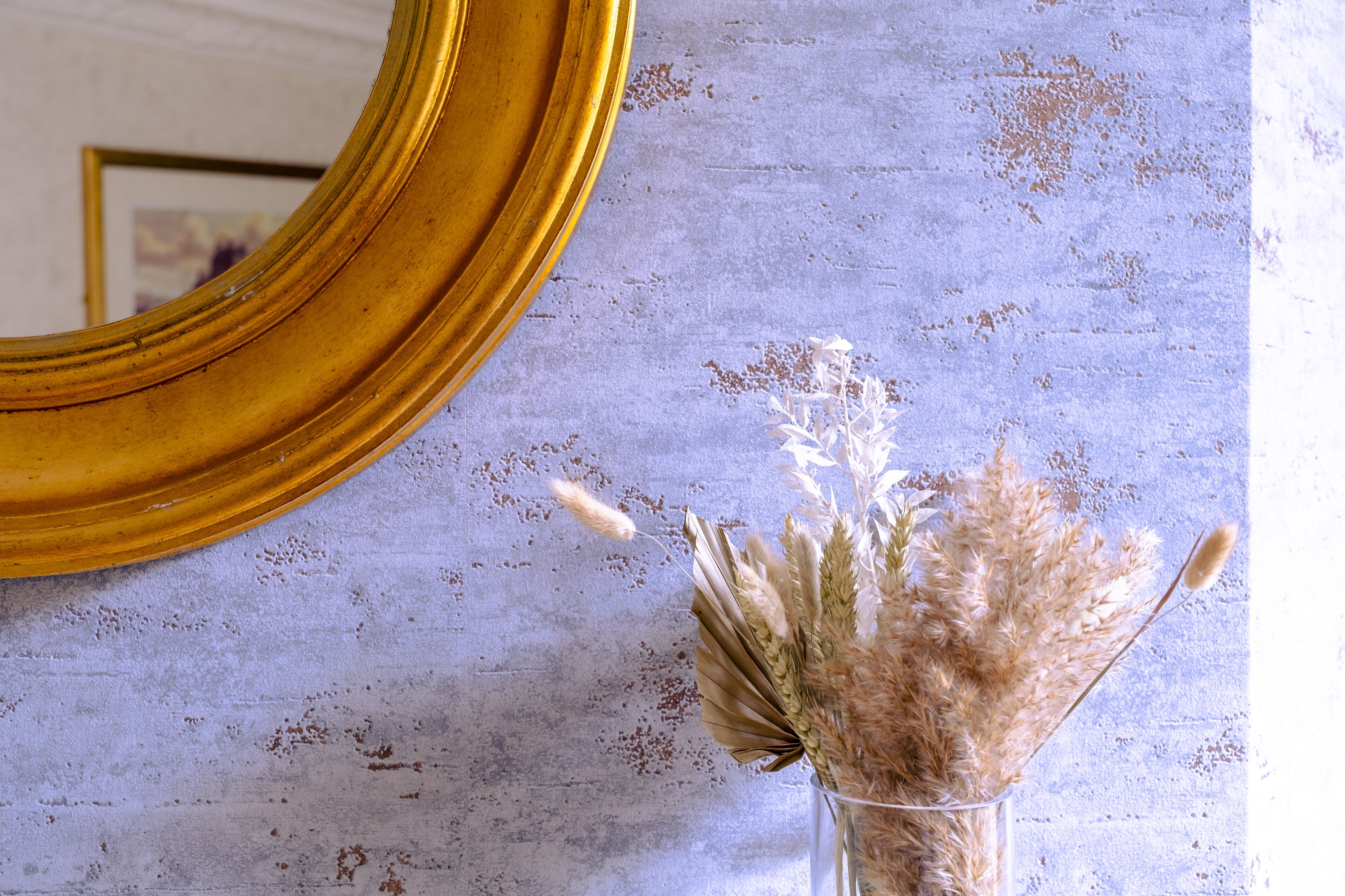A mirror with a gold frame, a vase with dried flowers, and a textured wall.