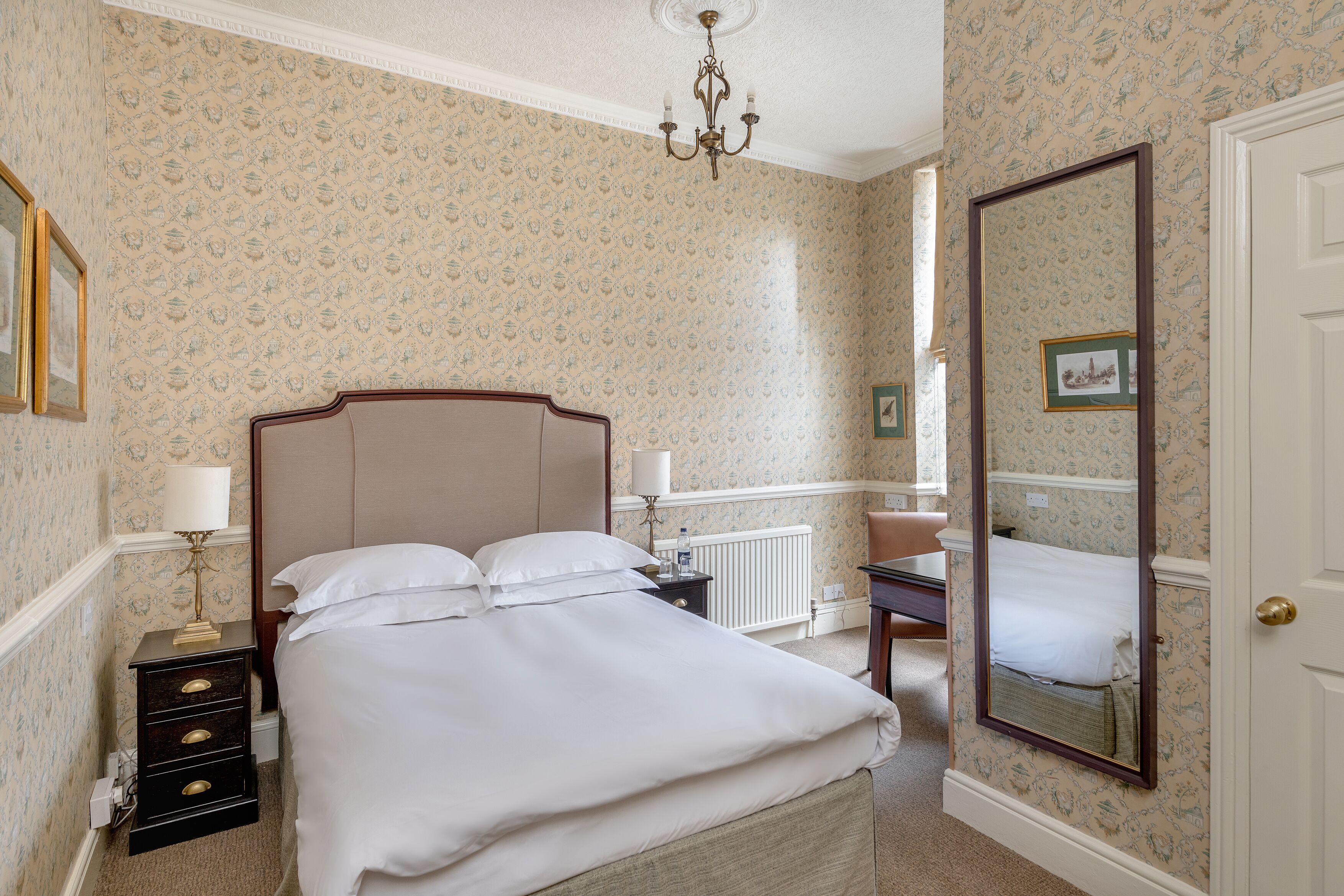 A neatly made bed with white linens, a bedside table with a lamp, and a large mirror on the wall.
