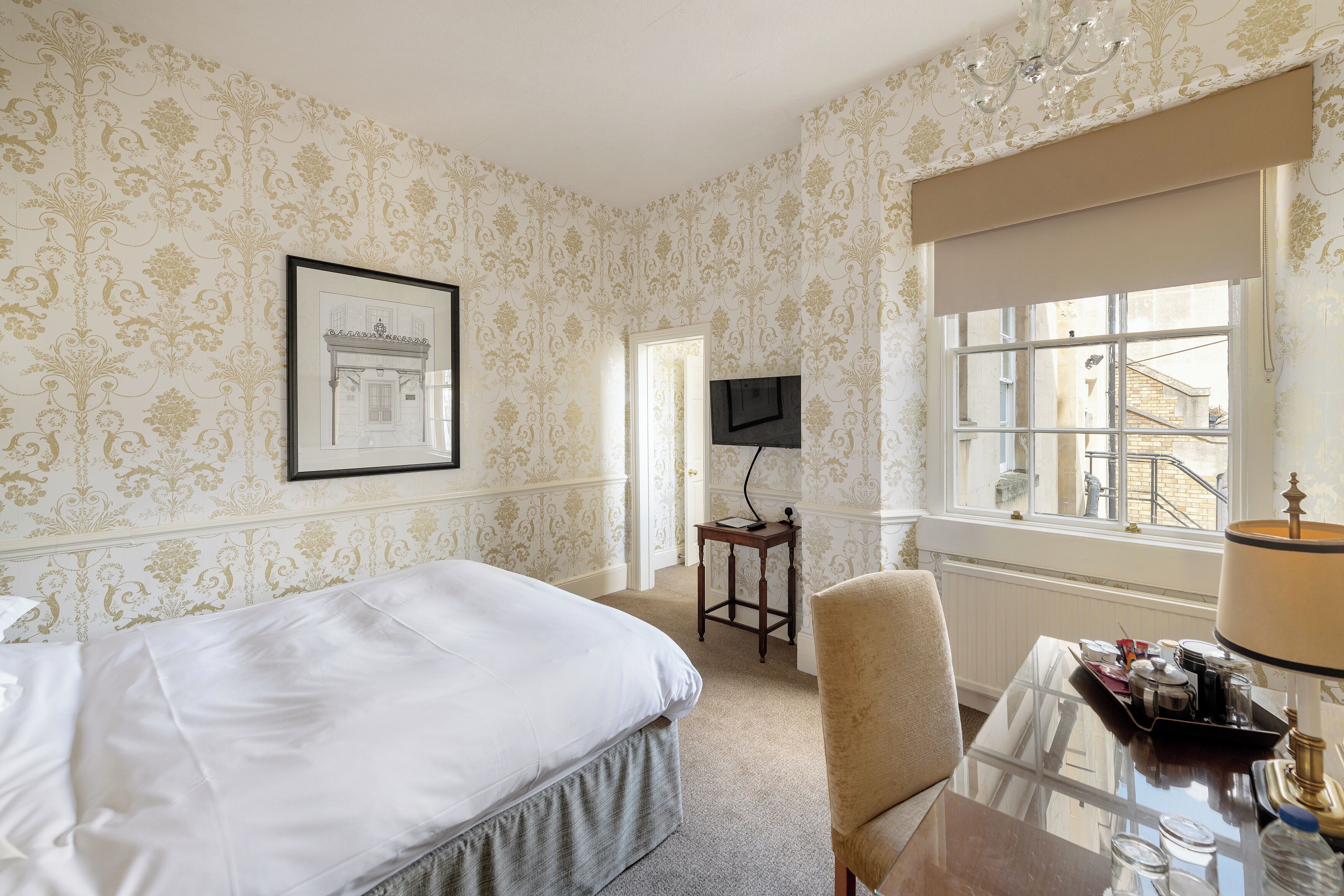 A hotel room with a bed, a desk with a lamp, a chair, a television, and a window with a view of buildings.
