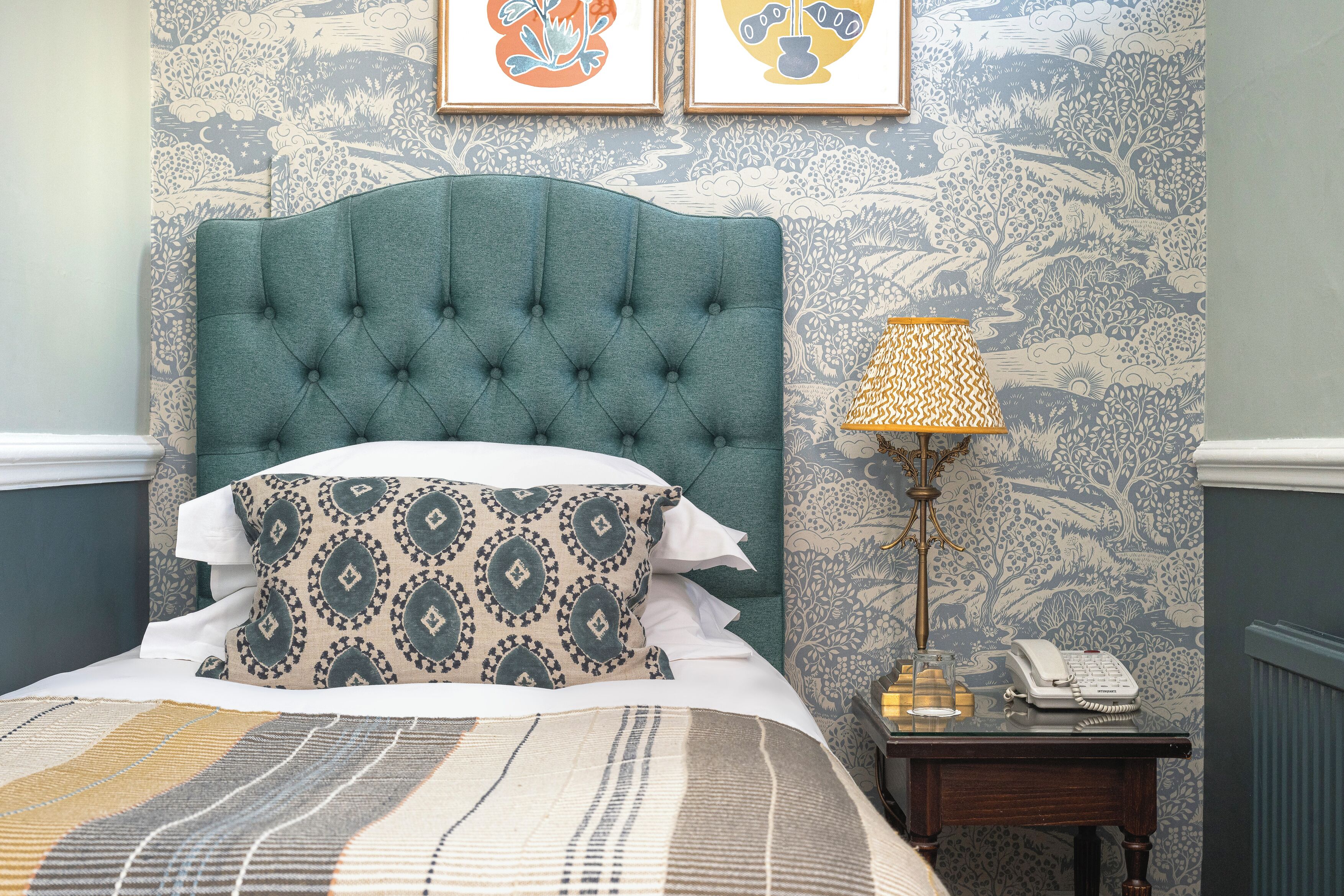 A neatly made bed with patterned pillows, a tufted headboard, and a bedside table with a lamp and a telephone.