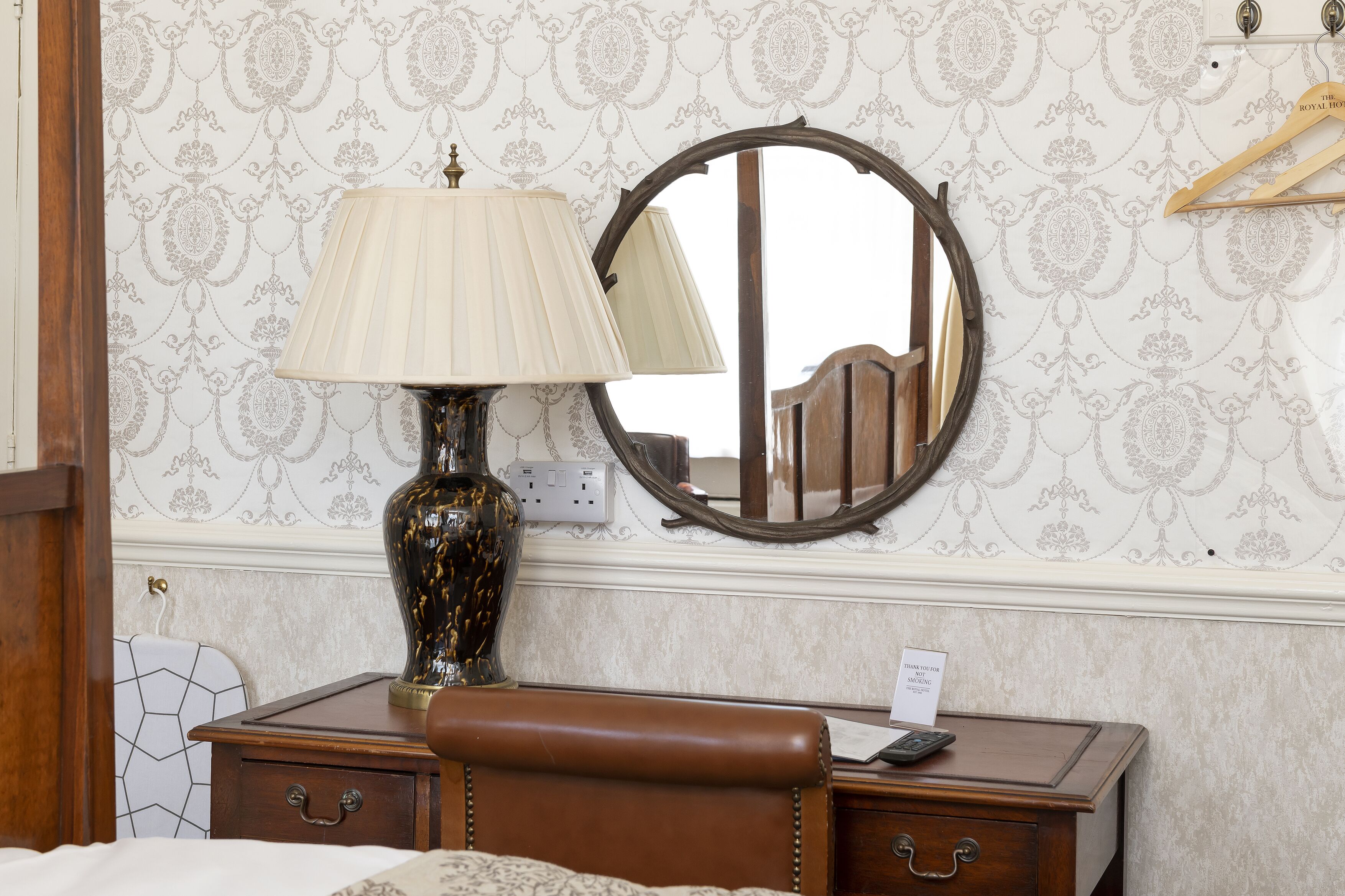 A hotel room with a wooden desk, a lamp, a mirror, and a sign indicating a room for sale.