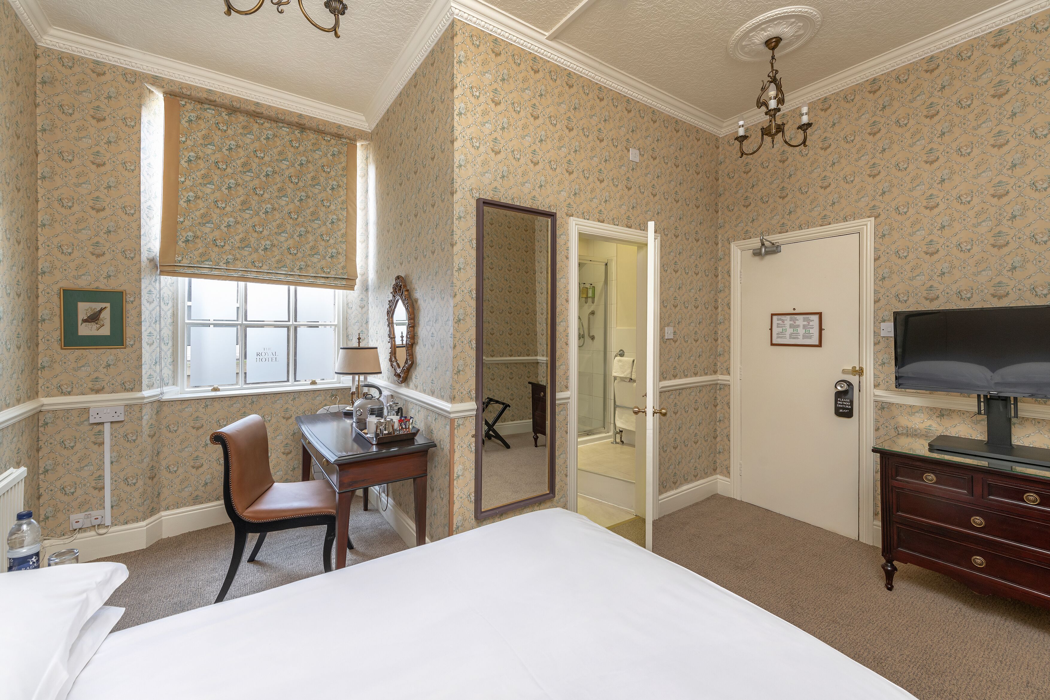 Quarto de hotel com cama, escrivaninha com cadeira, televisão e banheiro visível por uma porta aberta.